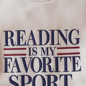 May include: Cream-colored sweatshirt with the embroidered phrase "READING IS MY FAVORITE SPORT" in navy blue lettering. Two red horizontal lines are embroidered above the words "IS MY". The sweatshirt has a crew neck.