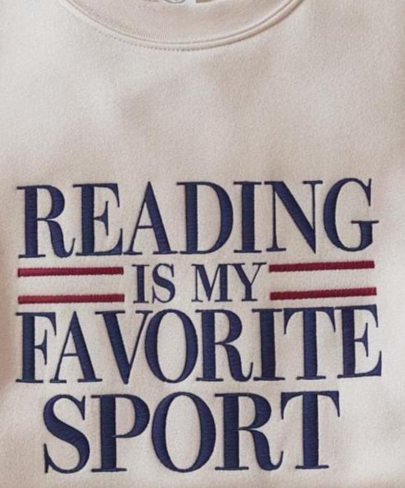 May include: Cream-colored sweatshirt with the embroidered phrase "READING IS MY FAVORITE SPORT" in navy blue lettering. Two red horizontal lines are embroidered above the words "IS MY". The sweatshirt has a crew neck.