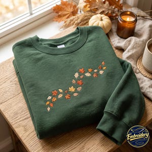Könnte beinhalten: Ein waldgrünes Sweatshirt mit Rundhalsausschnitt und einem gestickten S-förmigen Muster aus Herbstblättern in Orange-, Gelb-, Braun- und Cremetönen. Das Design ist mittig auf der Brust platziert und zeigt detaillierte Stickereien der Blätter.