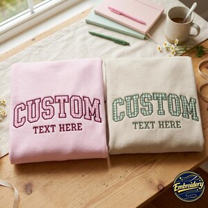 May include: Two folded sweatshirts, one light pink and one beige, each featuring embroidered collegiate-style lettering that reads CUSTOM TEXT HERE. The pink sweatshirt has pink gingham letters, while the beige sweatshirt has green gingham letters.