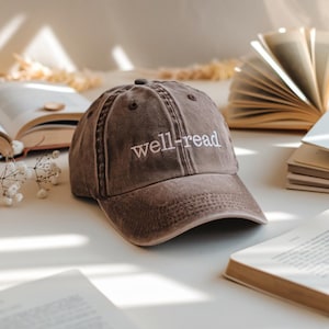 May include: A brown baseball cap with the embroidered text "well-read" in white. The cap has a worn appearance and is surrounded by open books, suggesting a literary theme. The cap has a curved peak and a classic six-panel design.