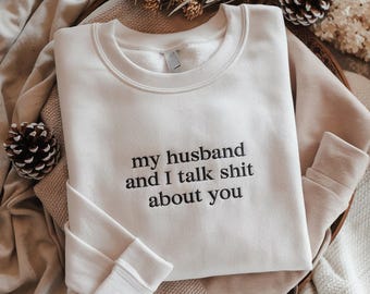 Może przedstawiać: Kremowa bluza z napisem "my husband and I talk shit about you" w kolorze czarnym. Bluza ma okrągły dekolt i długie rękawy ze ściągaczami. Odzież jest prezentowana na neutralnej powierzchni z elementami dekoracyjnymi.