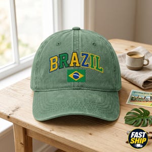 Peut inclure: Une casquette de baseball vert sauge arborant le mot BRAZIL brodé dans une police universitaire jaune, verte et bleue, placé au-dessus d'un petit écusson rectangulaire brodé du drapeau national du Brésil avec ses couleurs verte, jaune et bleue.