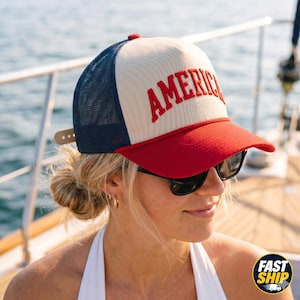 Könnte beinhalten: Eine Trucker-Cap mit cremefarbenem Vorderteil, auf dem das Wort AMERICA in Rot gestickt ist, einem roten Schirm und einem dunkelblauen Netzrücken. Die Kappe verfügt über ein rotes Kordeldetail an der Basis des Vorderteils und einen verstellbaren Verschluss.