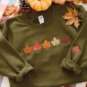 May include: An olive green sweatshirt featuring embroidered autumn leaves in shades of orange, red, and brown. A single leaf is embroidered on the sleeve. The sweatshirt is displayed on a plaid surface with autumn foliage and a small pumpkin.