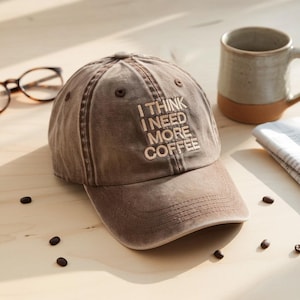 May include: A brown baseball cap with the embroidered text "I THINK I NEED MORE COFFEE." The cap is made of a worn, textured fabric. A ceramic mug and eyeglasses are in the background.