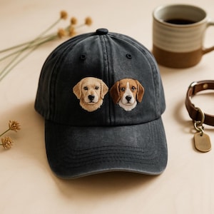 May include: A dark grey baseball cap with embroidered images of a Golden Retriever and a Beagle. The cap has a curved peak and a button on top. A brown leather dog collar and a coffee mug are in the background.