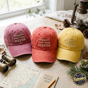 May include: Three baseball caps in dusty rose, terracotta, and mustard yellow, each featuring the embroidered white text "support your local cryptid" on the front panel. The caps have a structured crown, a curved brim, and eyelet ventilation.