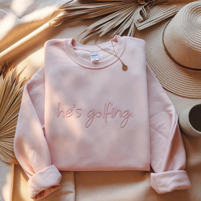 May include: A light pink sweatshirt with the embroidered phrase "he's golfing" in a cursive font. The long-sleeved top is folded, with a gold necklace and a straw hat in the background. The sweatshirt is a casual, comfortable garment.