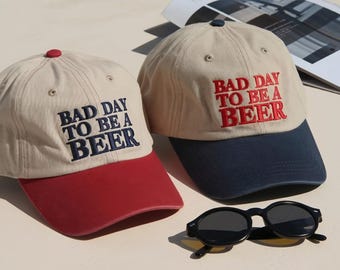 May include: Two beige baseball caps with embroidered text reading BAD DAY TO BE A BEER. One cap features a red brim and dark blue text, while the other has a dark blue brim and red text. A pair of black sunglasses rests in front of the caps.