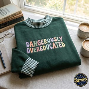 May include: A dark green sweatshirt featuring a mock neck collar and sleeve cuffs with a white and green gingham pattern. The chest displays the embroidered text DANGEROUSLY OVEREDUCATED in a multi-coloured, rounded, bubbly font style.