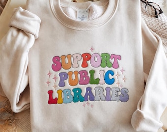 May include: A cream-colored crewneck sweatshirt featuring the embroidered text "SUPPORT PUBLIC LIBRARIES" in a multi-colored, bubbly font. The design is accented with small, embroidered pink star shapes scattered around the lettering.