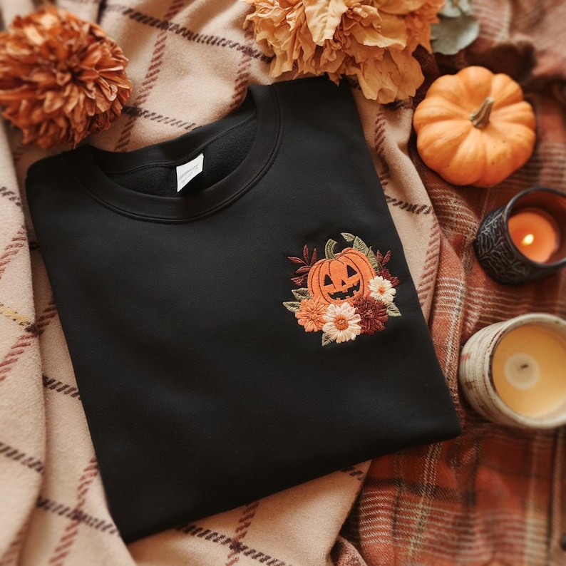 Peut inclure: Sweat-shirt noir avec une broderie de citrouille et de fleurs. La citrouille orange a un visage sculpt&eacute;, entour&eacute;e de fleurs orange, blanches et bordeaux. Le sweat-shirt est pos&eacute; sur une couverture &agrave; carreaux avec des d&eacute;corations d'automne.