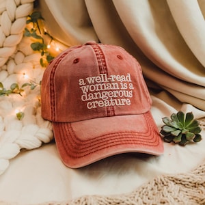May include: A red baseball cap with the text "a well-read woman is a dangerous creature" embroidered in white.