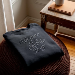 May include: A dark gray sweatshirt with the embroidered text "Woman of a Certain Rage" in a cursive font. The sweatshirt is folded and resting on a brown, textured ottoman. A small wooden table and a window are in the background.