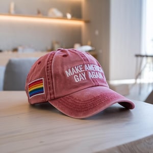 Può includere: Un berretto da baseball rosso con il testo "Make America Gay Again" ricamato in bianco. Una bandiera arcobaleno del pride è cucita sulla parte anteriore del berretto.