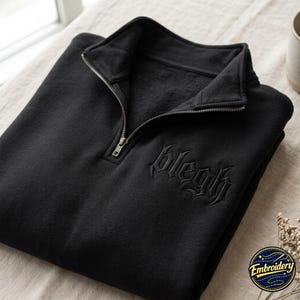 May include: A black quarter-zip sweatshirt featuring a silver-toned metal zipper and an embroidered design on the chest with the text "blegh" in a stylized gothic font. The garment has a high collar and a soft, fleece-like interior texture.