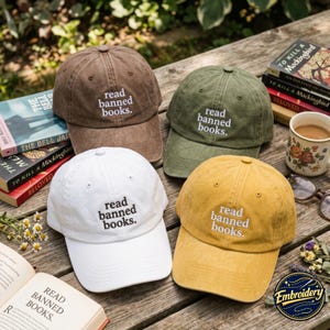 May include: Four baseball caps in brown, olive green, white, and mustard yellow, each featuring the embroidered text read banned books. The caps are arranged on a wooden surface alongside several books and a floral ceramic mug.