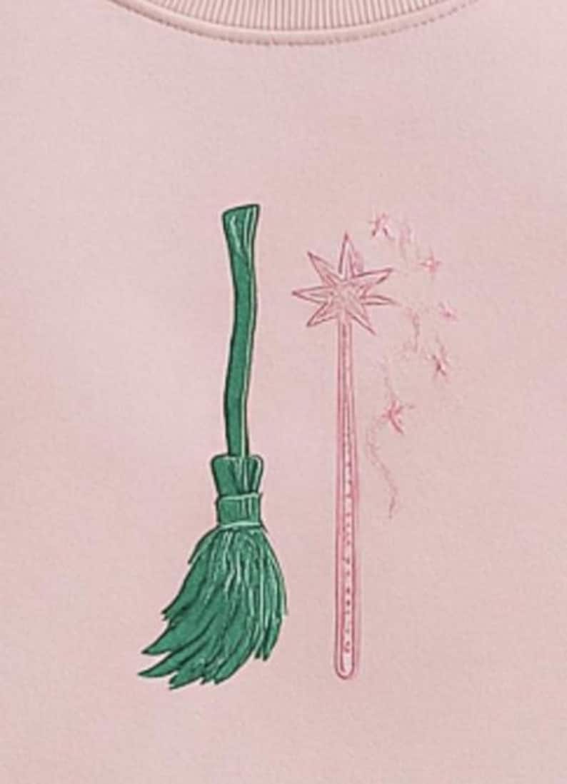 Op de afbeelding: Een lichtroze sweatshirt met geborduurde illustraties van een groene bezem en een roze toverstaf met steraccenten. Het ontwerp heeft een grillig, fantasiethema.