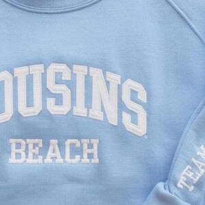 May include: Light blue sweatshirt with white embroidered text. The front of the sweatshirt reads "COUSINS BEACH" in a collegiate font. The sleeve features the text "TEAM CONRAD".