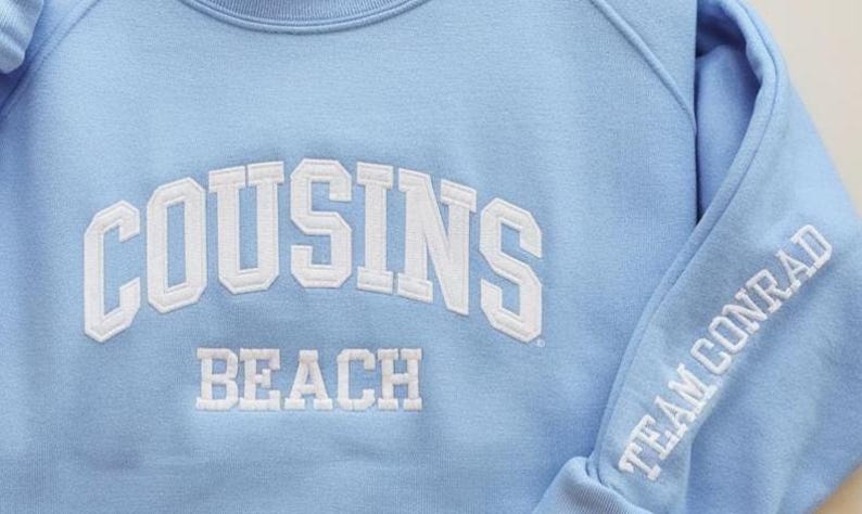 May include: Light blue sweatshirt with white embroidered text. The front of the sweatshirt reads "COUSINS BEACH" in a collegiate font. The sleeve features the text "TEAM CONRAD".