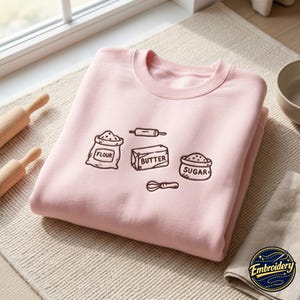 May include: A light pink crewneck sweatshirt featuring a dark brown embroidered graphic of baking ingredients and tools. The design includes a bag labeled Flour, a block labeled Butter, a bowl labeled Sugar, a rolling pin, and a wire whisk.