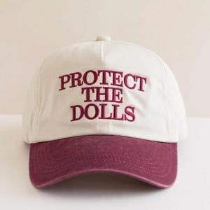 May include: A cream-colored baseball cap featuring the embroidered text "PROTECT THE DOLLS" in a deep maroon thread. The cap has a structured crown and a curved, solid maroon brim, with a matching button at the top center of the crown.