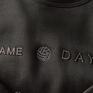 Może przedstawiać: Ciemnoszara bluza z napisem "GAME DAY" wyhaftowanym jaśniejszą szarą nicią. Pomiędzy słowami znajduje się grafika siatk&oacute;wki. Bluza ma okrągły dekolt i miękki, teksturowany wygląd.
