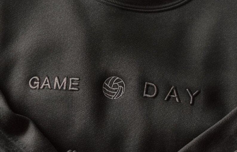 Może przedstawiać: Ciemnoszara bluza z napisem "GAME DAY" wyhaftowanym jaśniejszą szarą nicią. Pomiędzy słowami znajduje się grafika siatk&oacute;wki. Bluza ma okrągły dekolt i miękki, teksturowany wygląd.