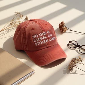 May include: A terracotta baseball cap with the slogan "NO ONE IS ILLEGAL ON STOLEN LAND" in white. The cap sits on a cream surface, alongside a notebook, spectacles, and dried flowers.