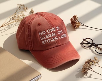 May include: A dusty rose baseball cap with the text "NO ONE IS ILLEGAL ON STOLEN LAND" in white. The cap is on a light beige surface with a notebook, eyeglasses, and dried flowers.