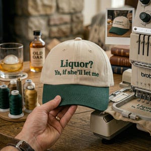 May include: A beige and green baseball cap with the text "Liquor? Ya, if she'll let me" embroidered in green. The cap has a curved brim and is being held up by a hand. The image also shows a glass of whiskey with an ice sphere, a bottle of Old Oak whiskey, and spools of thread.