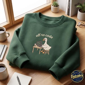 May include: A dark green crewneck sweatshirt featuring a graphic of a white goose wearing glasses, sitting at a small desk with a laptop and a coffee cup. The text above the goose reads "off to code" in a simple, light-colored sans-serif font.