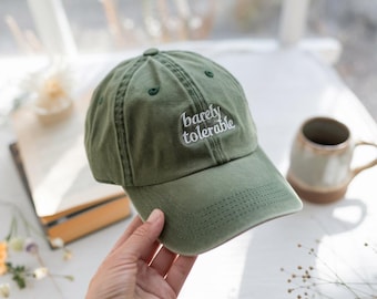 Puede incluir: Gorra de béisbol verde oliva con el texto bordado "barely tolerable" en blanco. La gorra tiene una visera curva y un cierre de correa de tela. Un accesorio casual.