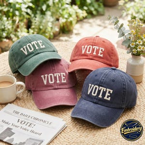 May include: Four baseball caps in sage green, dusty rose, terracotta, and navy blue, each featuring the word VOTE embroidered in white capital letters on the front. The caps have a distressed, washed finish and a curved brim with a button on top.
