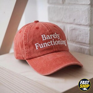 May include: A washed-out terracotta red baseball cap featuring the white embroidered text Barely Functioning on the front panel. The hat has a curved brim, a button on the top, and stitched eyelets for ventilation, showcasing a worn-in aesthetic.