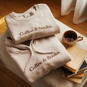 May include: Two beige sweatshirts, one crew neck and one hooded, both embroidered with "Coffee & books." A ceramic mug with a dark drink and a stack of books are next to the clothing. The hoodie has a drawstring.