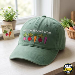 Puede incluir: Gorra de béisbol verde con la frase bordada "Let's root for each other" e ilustraciones de verduras coloridas. La gorra tiene una visera curva y un botón en la parte superior. Un paquete de semillas de verduras es visible en el fondo.
