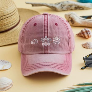 May include: A faded rose baseball cap with a curved peak. The cap has embroidered white designs of waves, a turtle, and a sun. The cap is made of a soft, faded fabric, and has a classic six-panel design.