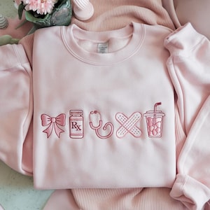 May include: A light pink sweatshirt with embroidered medical-themed designs. The designs include a bow, prescription bottle, stethoscope, crossed bandages, and a drink with a straw. The sweatshirt is made of a soft material.