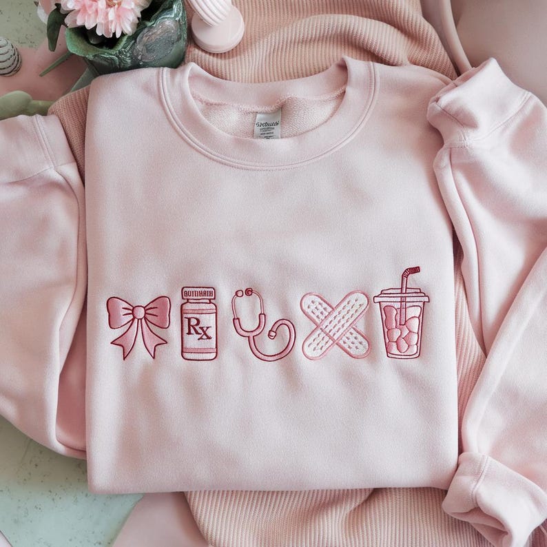 May include: A light pink sweatshirt with embroidered medical-themed designs. The designs include a bow, prescription bottle, stethoscope, crossed bandages, and a drink with a straw. The sweatshirt is made of a soft material.