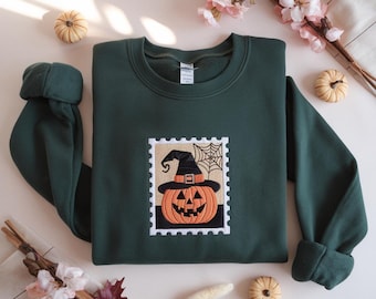 May include: Dark green sweatshirt with a Halloween patch. The patch is a postage stamp design featuring an orange jack-o'-lantern wearing a witch's hat and a spider web. The sweatshirt has long sleeves and a crew neck.