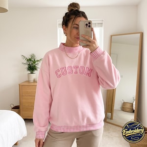 May include: A light pink sweatshirt featuring a mock neck collar, sleeve cuffs, and a bottom hem all detailed with a pink and white gingham pattern. The word CUSTOM is embroidered across the chest in a pink and white gingham fabric applique.