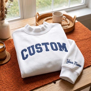 May include: A white long-sleeve sweatshirt featuring a mock neck collar and ribbed cuffs. The word CUSTOM is embroidered in large navy blue block letters across the chest, while the phrase Your Name is embroidered in navy blue on the sleeve cuff.