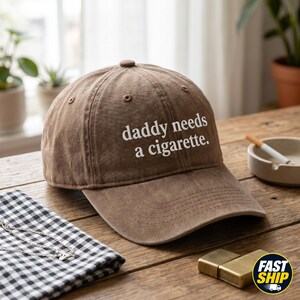 Op de afbeelding: Bruine baseballpet met de tekst "daddy needs a cigarette." De pet is gemaakt van een zacht materiaal en heeft een gebogen klep. Een sigaret en aansteker zijn zichtbaar op een houten tafel.