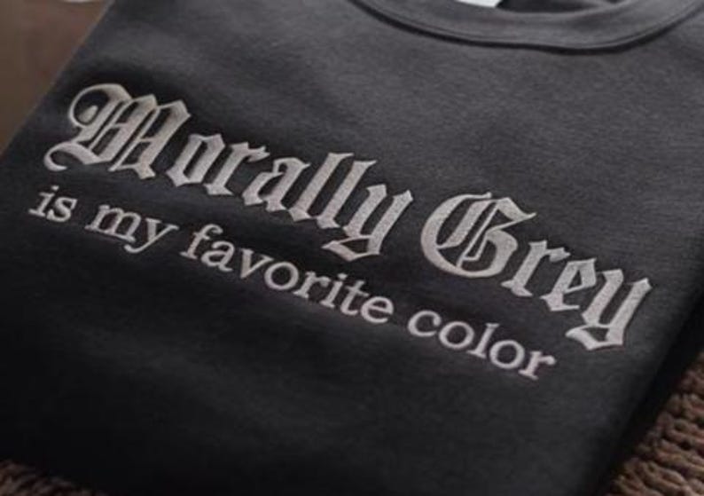 以下が含まれることがあります： ゴシック体で「Morally Grey is my favorite color」と刺繍されたダークグレーのスウェットシャツ。 文字は明るいグレーで、生地とのコントラストを生み出しています。
