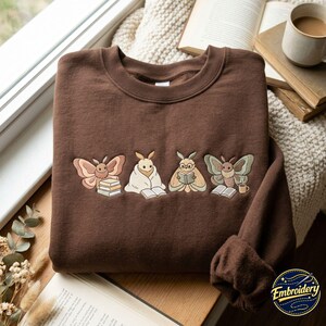 May include: A dark brown crewneck sweatshirt featuring an embroidered design of four moths engaged in reading activities. The moths are depicted with books, a blanket, and a small mug, rendered in soft pastel tones of beige, pink, and green.