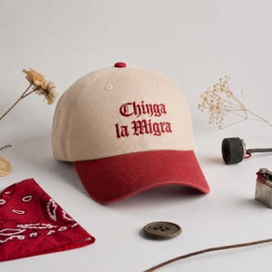 Può includere: Un cappellino da baseball beige con visiera rossa e un bottone rosso sulla parte superiore. La parte anteriore del cappellino presenta il testo ricamato "Chinga la Migra" in un carattere gotico rosso scuro. Il cappellino ha una corona.