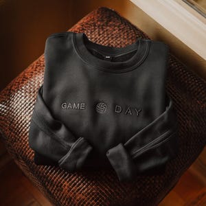 Może przedstawiać: Czarna bluza z napisem "GAME DAY" wyhaftowanym na piersi. Projekt zawiera grafikę siatk&oacute;wki między słowami. Bluza jest złożona i spoczywa na brązowej, teksturowanej powierzchni.