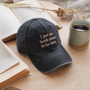 May include: A black baseball cap with the text "I put my book down to be here." embroidered in a light brown color.